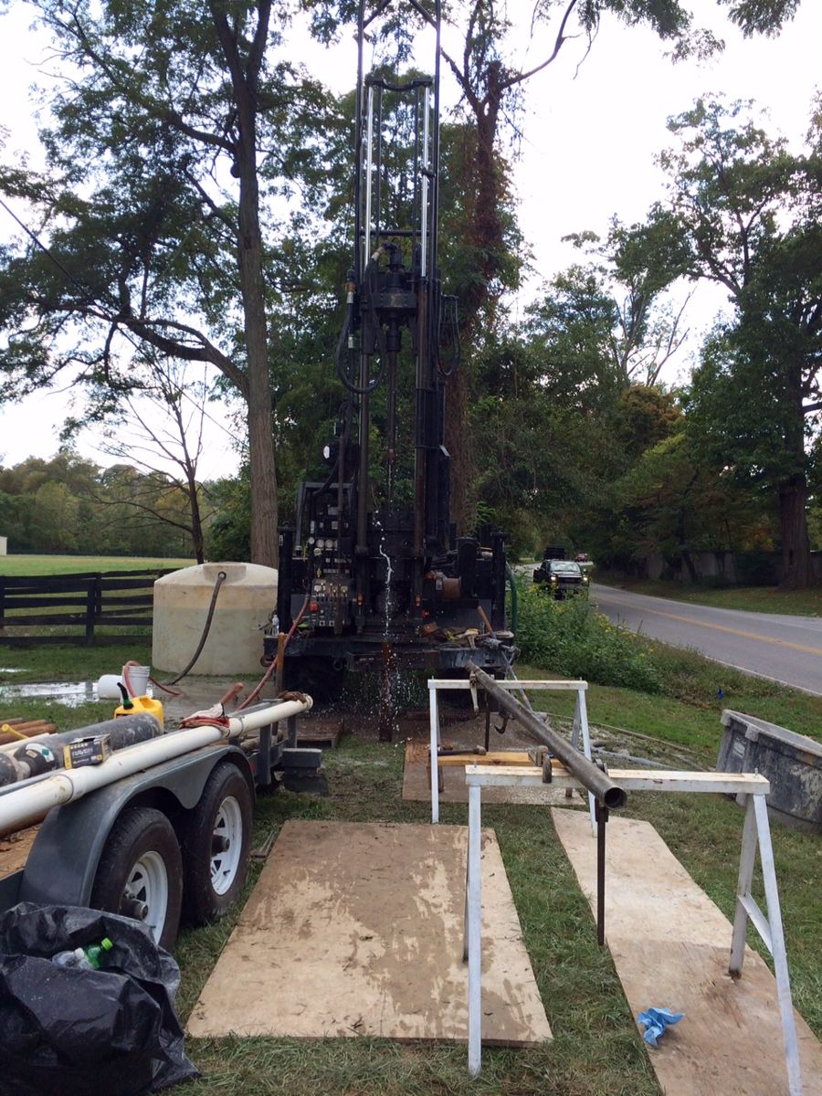 Truck-mounted drilling rig with its mast raised on a grassy roadside site