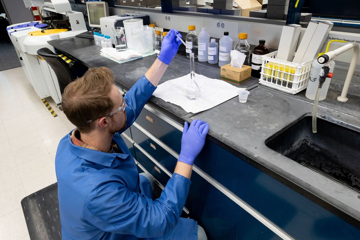 A chemist in a lab coat pipetting a water sample into a volumetric flask at a lab bench