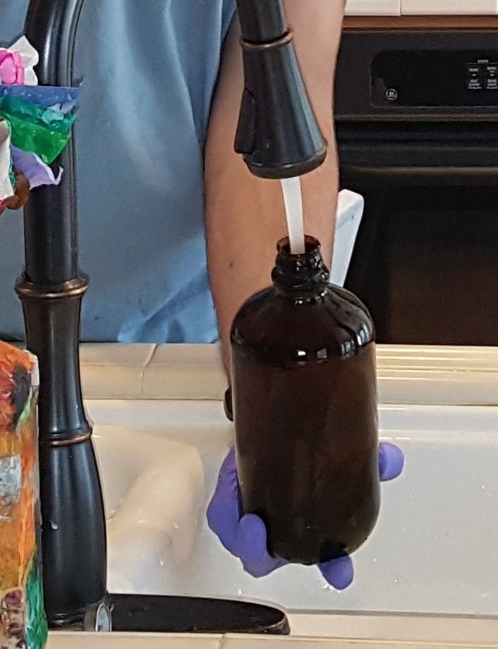 A gloved hand filling an amber glass sample bottle at a kitchen faucet
